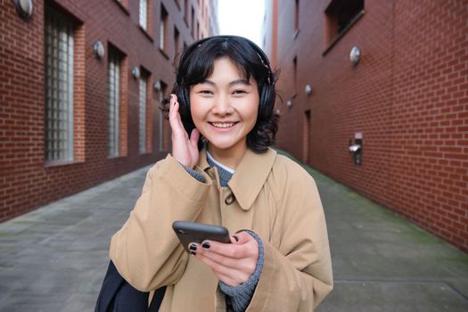 Beautiful smiling woman walking around town, listening music and using mobile phone app, going to college, wearing headphones.