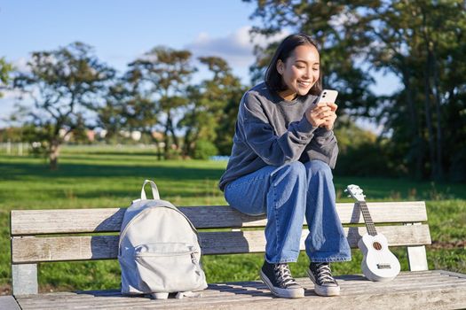 Young woman sitting in park on bench with ukulele, looking at smartphone, reading message on mobile phone and smiling.