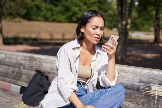 Asian girl frowning, looking concerned at her mobile phone, reading sad message, bad news on smartphone app, sitting on bench in park worrying.