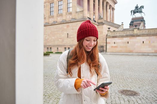 Tourism and sightseeing concept. Young redhead woman, tourist walks around city, looks at her smartphone app and at history stand, explores adventures.