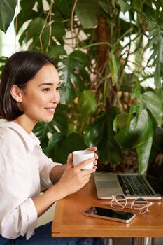 Vertical shot of beautiful woman sits in cafe with laptop, works or studies online at co-working space, smiling relaxed.