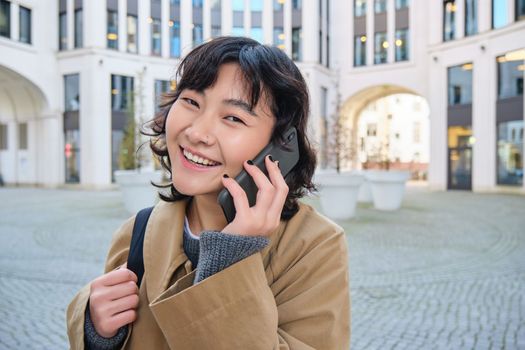 Portrait of smiling asian girl talks on mobile phone, speaks with friend on smartphone, walks in city centre, stands on street and laughs.