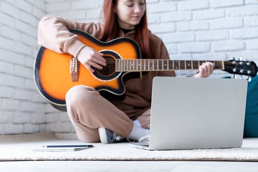 young caucasian red-haired woman sitting on rug in front of laptop and playing acoustic guitar at home
