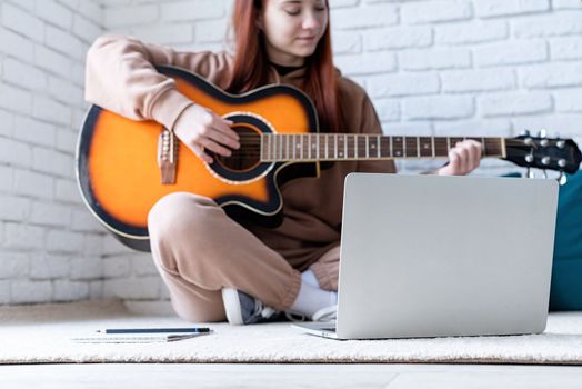 young caucasian red-haired woman sitting on rug in front of laptop and playing acoustic guitar at home