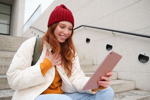 Happy stylish redhead girl, student in red hat, holds digital tablet, uses social media app, searches something online, connects to wifi.