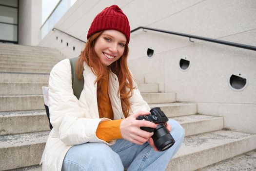 Smiling redhead girl photographer, checks her shots, holds camera and looks at screen, takes photos outdoors, walks around street and does streetstyle shooting.