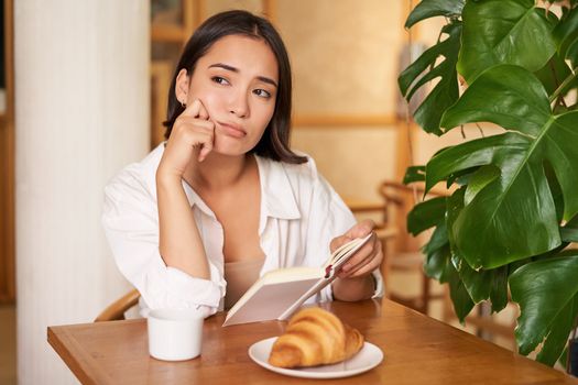 Young woman feels uneasy, reads sad book and sits in cafe with cup of coffee, looks around with sulking unhappy face.