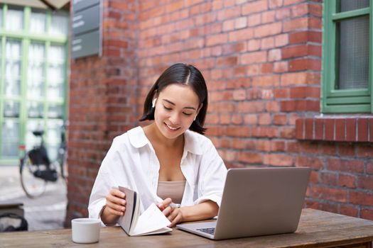 Working woman sitting in coworking space, drinking coffee and using laptop, wearing wireless headphones, watching video and smiling.