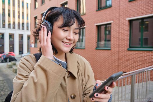 Young happy girl tourist, listens music in headphones, drinks takeaway and checks mobile phone, stands on street and smiles.