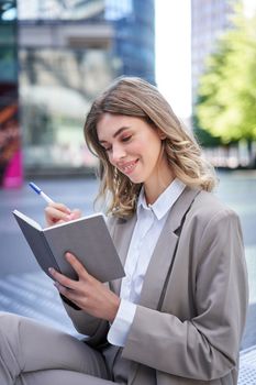 A young woman in corporate suit, sits with notebook and pen, takes notes, works and writes down her ideas.