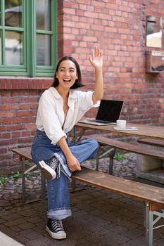 Vertical shot of happy girl attracting attention, waving hand at friend in cafe, sitting on bench with laptop.