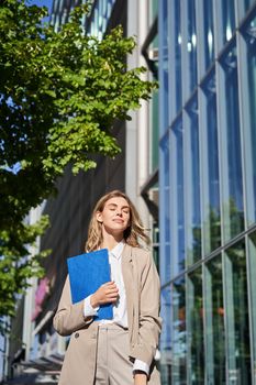 Portrait of confident saleswoman in her 30s, posing near office building on street, wearing beige suit, holding folder with documents.