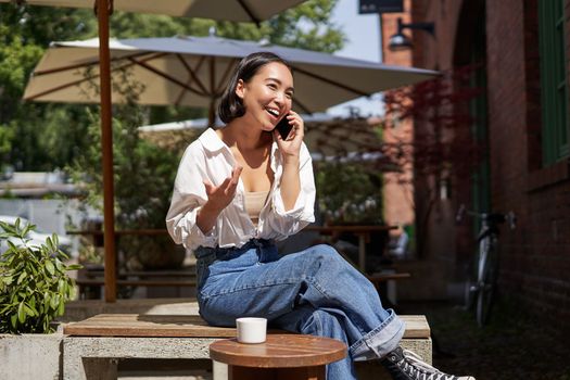 Beautiful young asian woman sitting in cafe with cup of coffee, talking on mobile phone, having lively conversation.