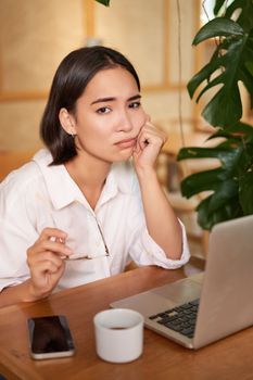Vertical shot of sad and gloomy young woman, sitting with laptop in cafe, grimacing and looking upset, feeling tired or disappointed.