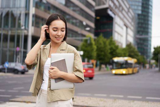 Portrait of beautiful asian woman standing on street with laptop and notebook, going to university or work, commute route.