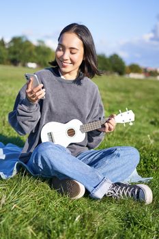 Vertical shot of smiling asian girl with smartphone, playing ukulele, reading chords or lyrics while singing, relaxing outdoors. Lifestyle and people concept.