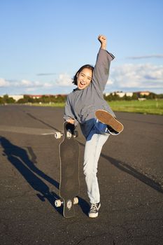 Vertical shot of asian girl feeling excited, skating on longboard, jumping and posing with skateboard, standing with cruiser on empty road, having fun outdoors.
