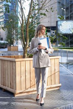 Beautiful businesswoman in suit, drinking takeaway coffee on street, waiting for someone near office building on lunch break.