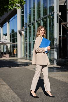 Portrait of businesswoman in beige suit and high heels walking on street. Corporate woman going to work, holding folder with documents.