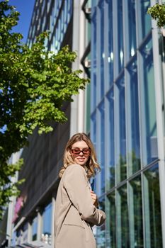Portrait of confident office lady, business woman in sunglasses posing outdoors, near her company office building, wearing beige suit.