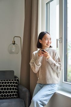 Portrait of young asian woman sitting near window and looking outside, drinking hot coffee from espresso cup and enjoying her cozy day off at home.