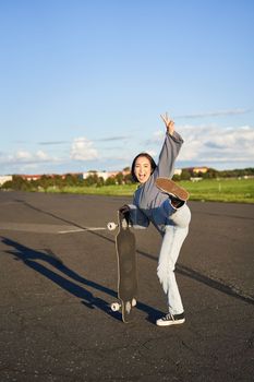 Vertical portrait of smiling asian woman standing on road with longboard, skateboarding on long cruiser, posing on empty road on sunny day.