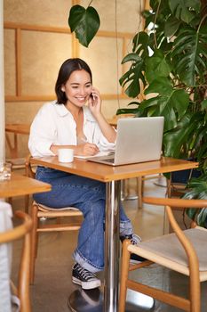 Stylish young woman answer phone call, sitting in cafe with laptop, working and taling on smartphone.