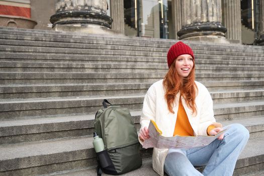 Portrait of young redhead woman, tourist sits with paper map and looks for a route to tourism attraction, rests on stairs outdoors.