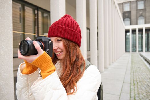 Smiling redhead girl photographer, taking pictures in city, makes photos outdoors on professional camera. Young talent and hobby concept