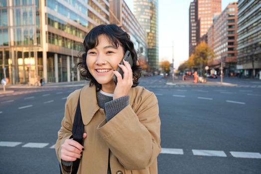 Smiling asian girl makes a phone call, stands on an empty street, calling someone on telephone, waiting for friend in city, going to a meeting.