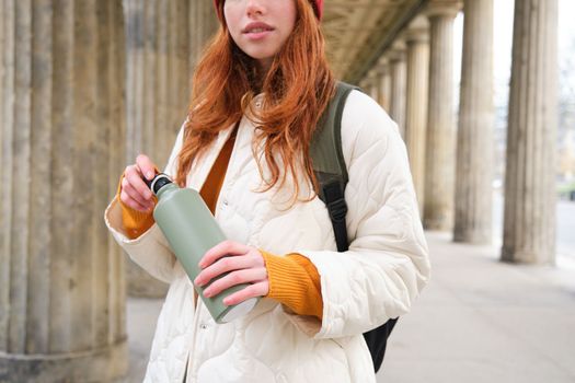 Close up portrait of redhead girl, tourist stands on street, opens thermos with hot drink, rests and pours herself coffee from flask.