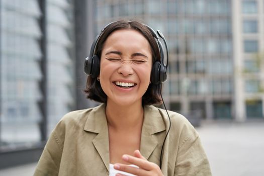 Carefree asian girl, laughing and smiling, wearing headphones and walking on street. Outdoor shot of young woman listening music and looking happy.