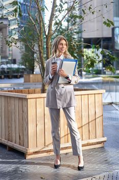 Stylish corporate woman in suit, standing on street with documents, work laptop and coffee, waiting near business center.