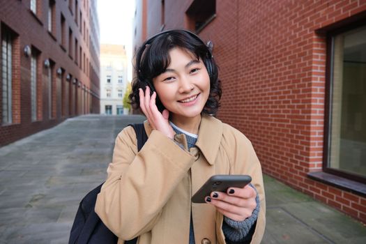 Portrait of young korean woman walking around city with backpack and headphones, listens music, looks at smartphone, uses phone application on streets.