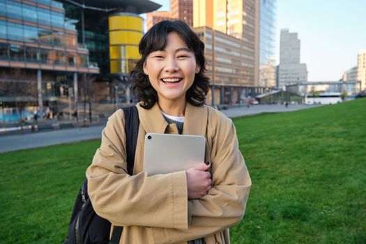 Image of korean girl with happy face, walks around town with student tablet, stands on street and smiles. Copy space