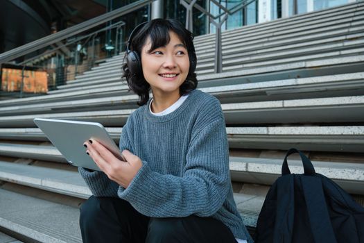 Smiling chinese student sits on stairs with tablet, draws digital art, graphic design project for freelance job, listens music in headphones and smiles.