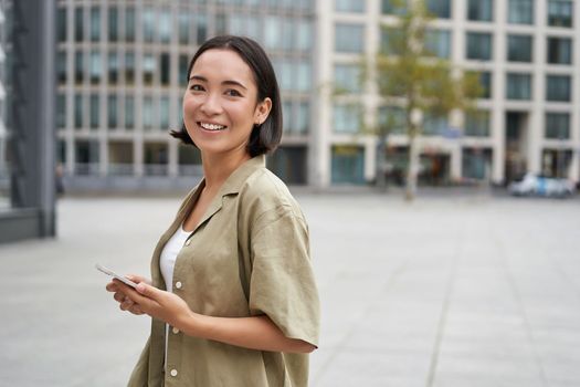 Portrait of asian woman standing on street, city square and holding mobile phone. Girl with smartphone walking outdoors.