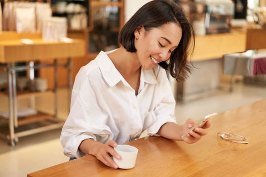 Portrait of beautiful asian woman with smartphone, relaxing in cafe, sitting and enjoying coffee while using mobile phone.