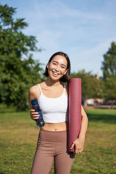 Portrait of young slim and healthy korean girl doing workout in park, standing with water bottle and rubber mat for execises on green lawn, smiling happily.