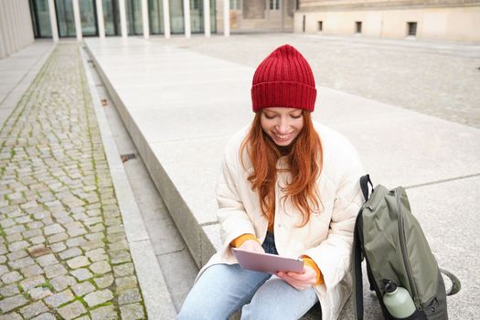 Beautiful redhead woman in red hat, sits with backpack and thermos, using digital tablet outdoors, connects to wifi, texts message, books tickets online.