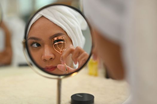 Reflecting in mirror of woman with towel on head using eyelash curler curling eyelashes. Beauty care and Cosmetics concept.