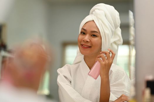 Beautiful young woman with bottle of perfume standing front of mirror. Beauty care and Cosmetics concept.