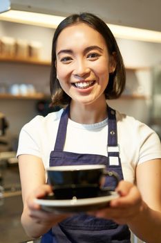 Portrait of smiling asian woman in apron, barista giving you cup of coffee, working in cafe, serving drinks.