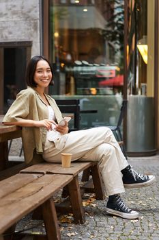 Portrait of stylish young korean woman sits in cafe, holds smartphone, smiles, enjoys coffee outdoors.