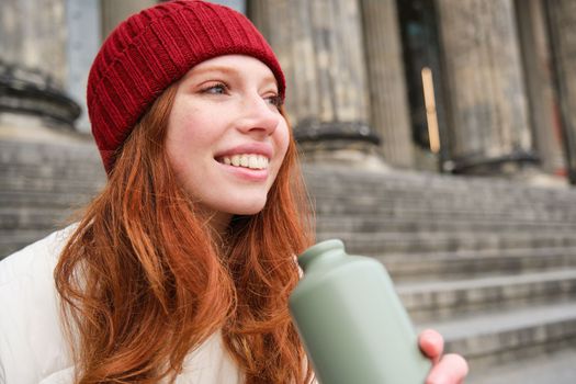Smiling redhead girl in red hat, sits on stairs near building with flask, drinks water and looks happy.