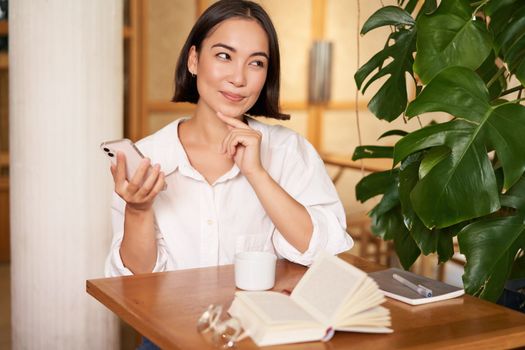 Beautiful young woman in cafe with cup of coffee and book, holding smartphone, using mobile phone, paying contactless waiting for waiter.