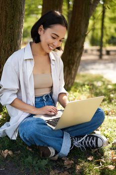 Beautiful asian girl sitting in park with laptop, working on remote, typing on keyboard, smiling at camera, resting beside tree.