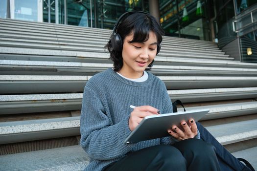 Young smiling digital artists works on her project using tablet and graphic pen, listens music in headphones and sits on stairs in public space, works on project.