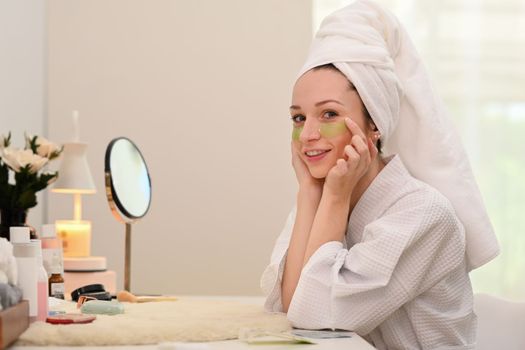 Portrait of caucasian woman in bathrobe applying anti fatigue under eyes. Beauty treatment and self care concept.