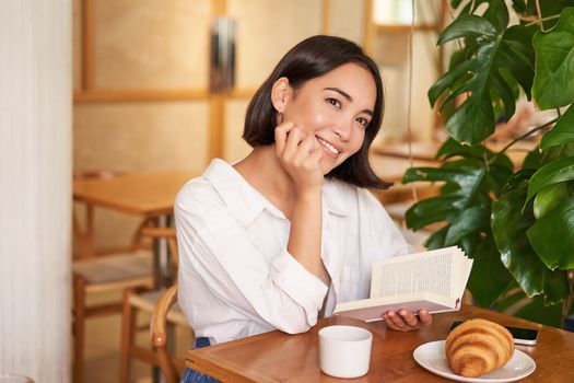 Romantic asian woman sitting with book in cafe, eating croissant and drinking coffee, reading and smiling, enjoying alone time.